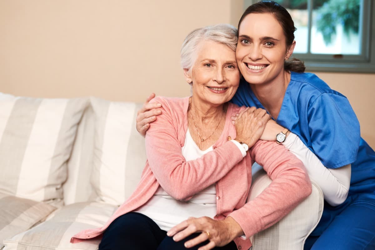 Cuidadora y mujer mayor abrazadas y sonrientes en el sofá de casa